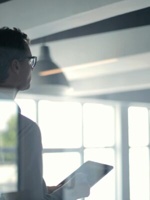 A businessman holding a tablet in an office, looking thoughtfully through a glass wall.