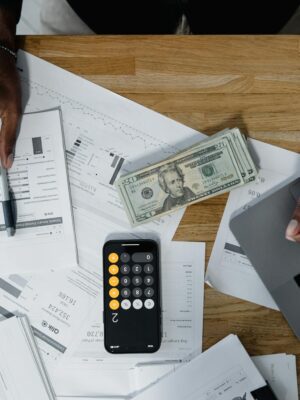Hands calculate finances with papers, cash, and a laptop on a wooden desk.