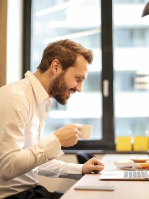 Man in office with coffee, smiling while working at laptop, captures the essence of remote work.
